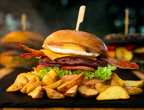 Hamburger z chrupiącym bekonem, świeżą sałatą i dojrzałym pomidorem, podany z grillowanymi ziemniakami na eleganckim czarnym talerzu.